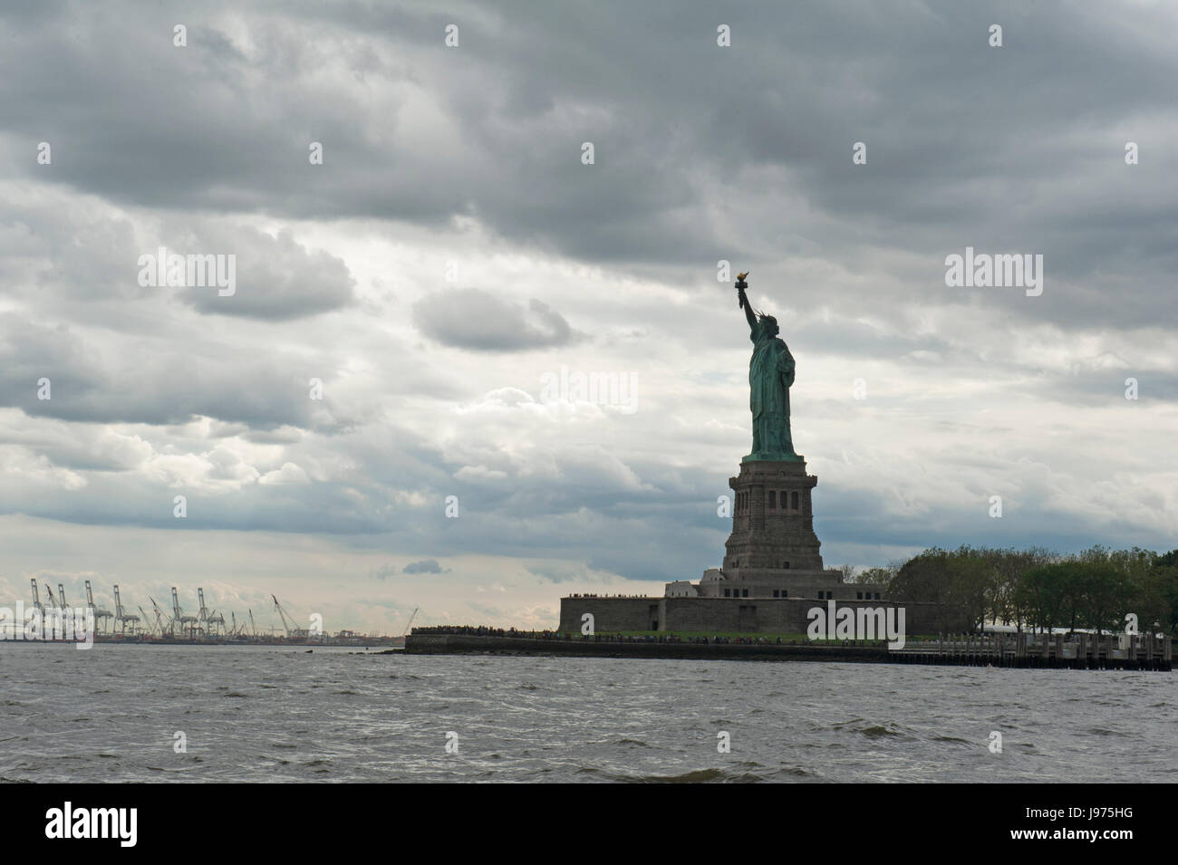 Die Statue of Liberty auf Liberty Island im New Yorker Hafen an einem bewölkten Tag in der Dämmerung mit die Gantries von Ports Elizabeth und Newark im Hintergrund. Stockfoto