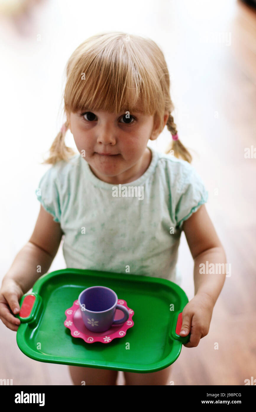 Kleine blonde Mädchen, Kind mit Fransen und Pigtails Holding ein Tablett mit einer Teetasse mit Untertasse, spielen Tee mit ihrer Puppe, kid Kinder Spielzeug Stockfoto