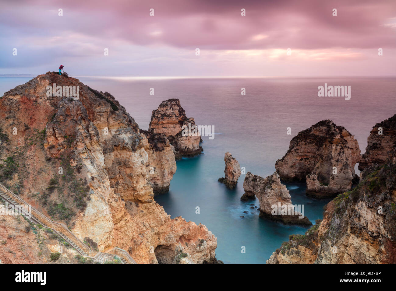 Fotograf auf Klippen umgeben von Meer unter dem rosa Himmel bei Sonnenaufgang, Ponta Da Piedade, Lagos, Algarve, Portugal Stockfoto