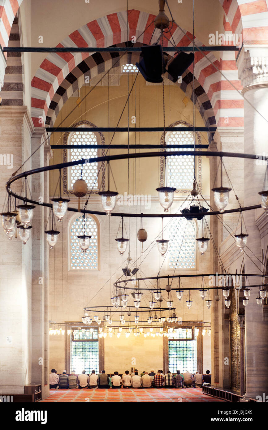 Die Menschen saßen im Gebet in einer historischen Moschee und zeigten kunstvolle Bögen, Hängelampen und islamische Architektur unter sanfter Beleuchtung. Stockfoto