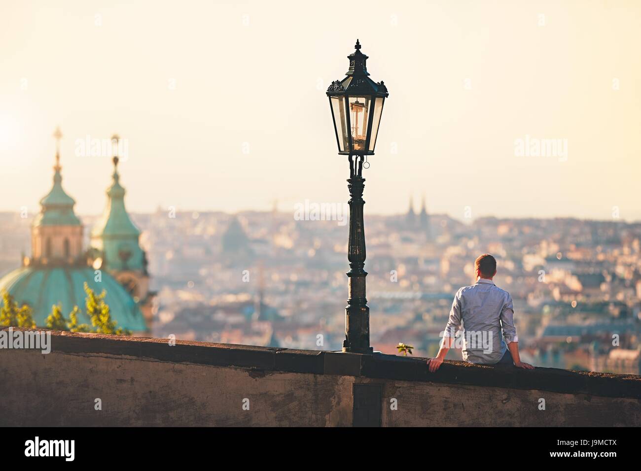 Junger Mann auf der Mauer sitzend und Skyline der Stadt bei Sonnenaufgang zu beobachten. Prag, Tschechische Republik Stockfoto