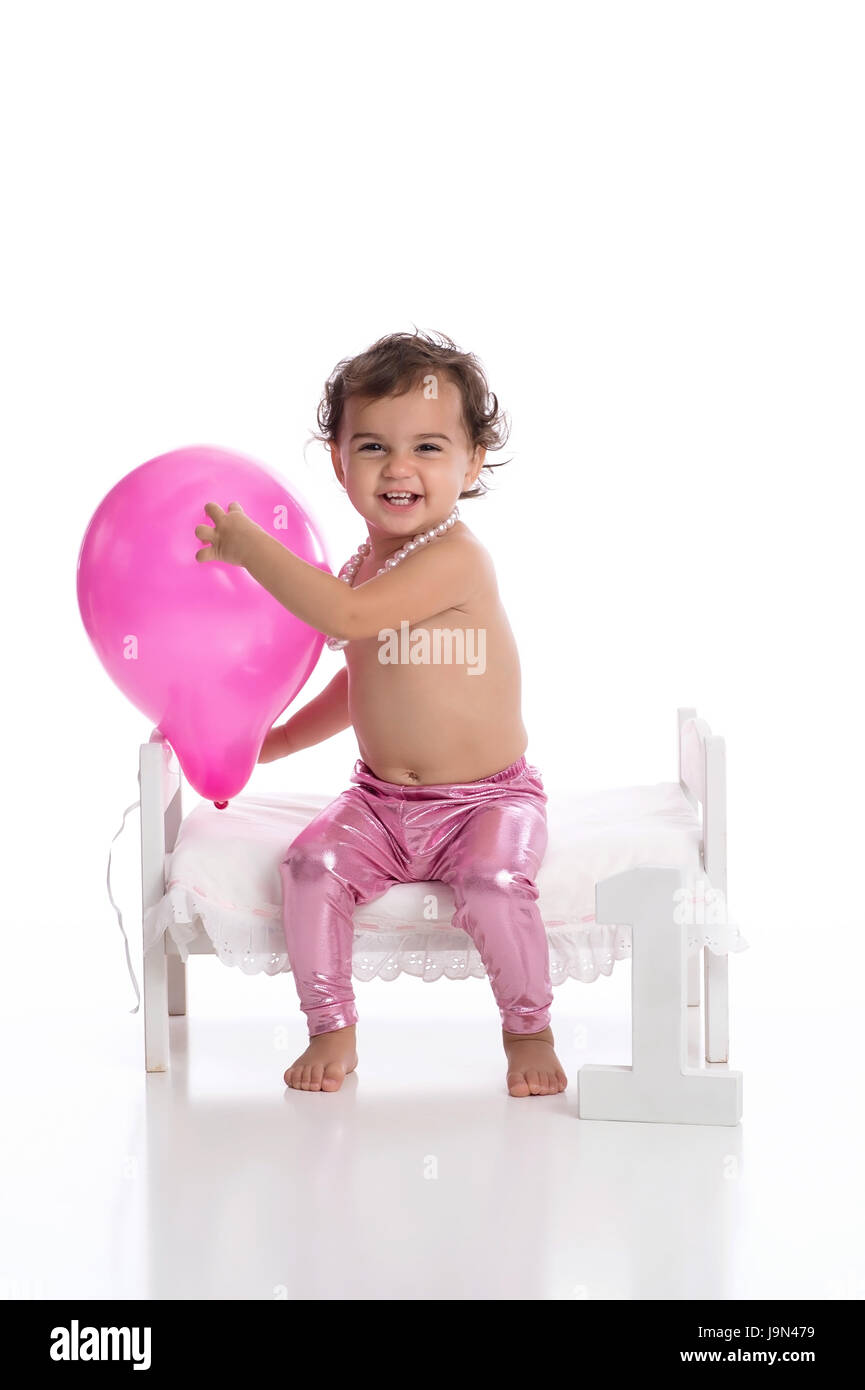 Ein Lachen, ein Jahr alt, Baby Mädchen Rosa Ballon halten. Sie trägt metallic rosa Leggins, eine Zeichenkette der Perlen und sitzen auf einem kleinen Bett auf einem w Stockfoto