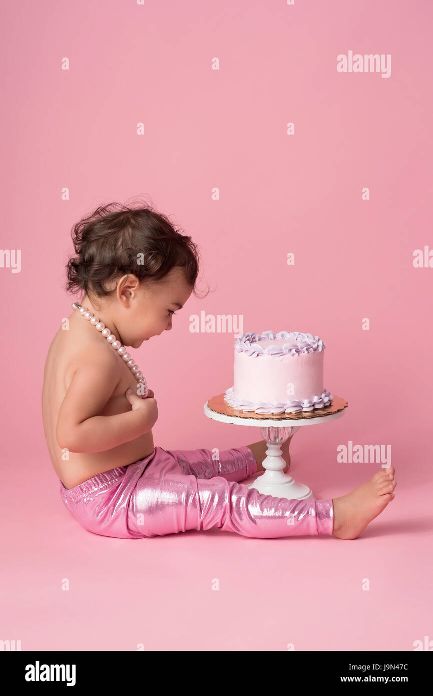 Ein Höhenplan der ein einjähriges Mädchen sitzt mit einem Kuchen. Sie trägt rosa Leggins und eine Zeichenkette der Perlen. Stockfoto