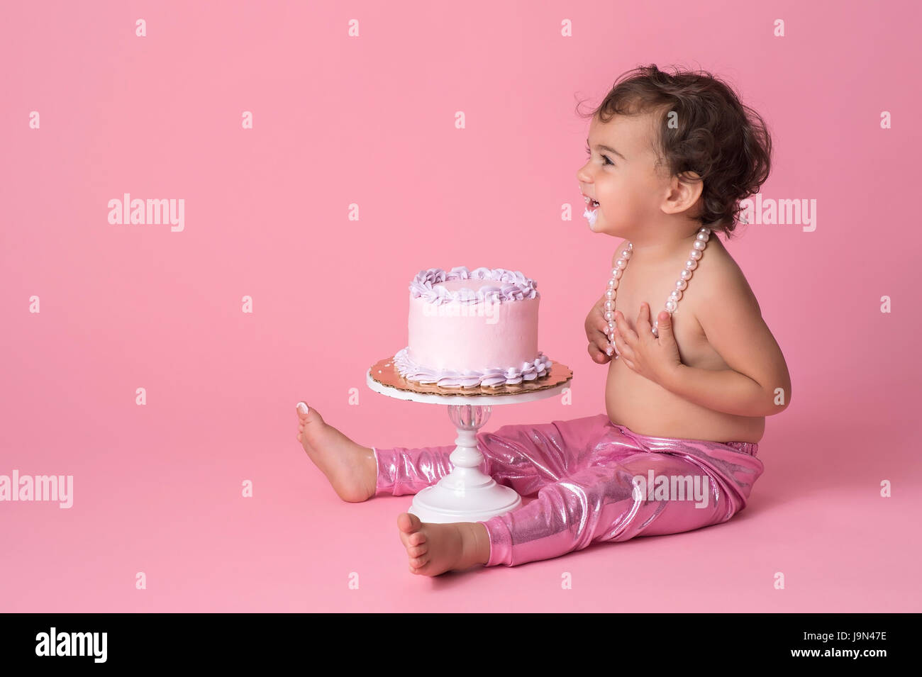Ein glückliches, ein Jahr alt, Baby Mädchen sitzt mit einem Kuchen. Sie trägt rosa Leggins, eine Zeichenkette der Perlen und Zuckerguss am Kinn hat. Stockfoto