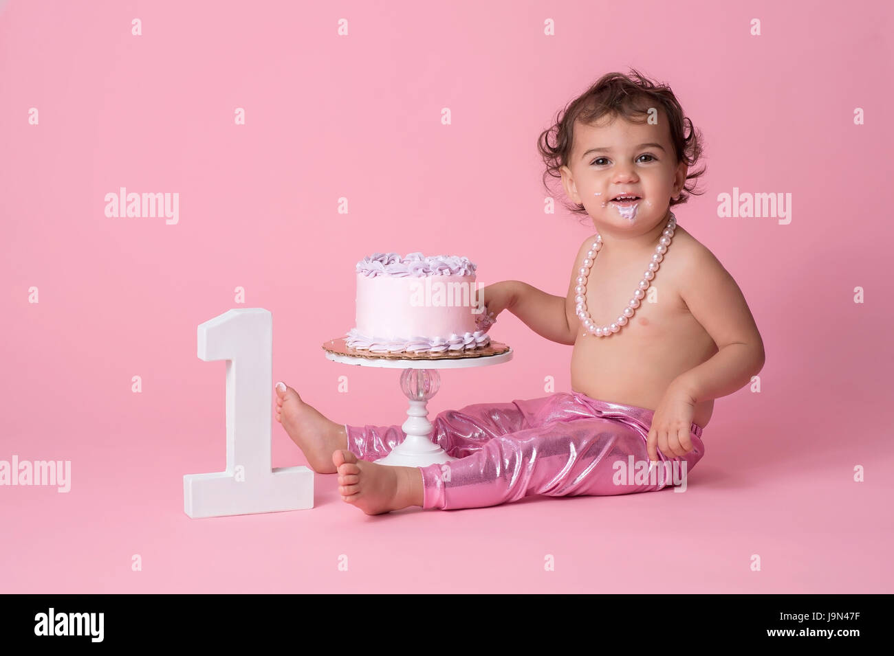 Ein glückliches, ein Jahr alt, Baby Mädchen sitzt mit einer Geburtstagstorte. Sie trägt rosa Leggins, eine Zeichenkette der Perlen und Zuckerguss am Kinn hat. Stockfoto