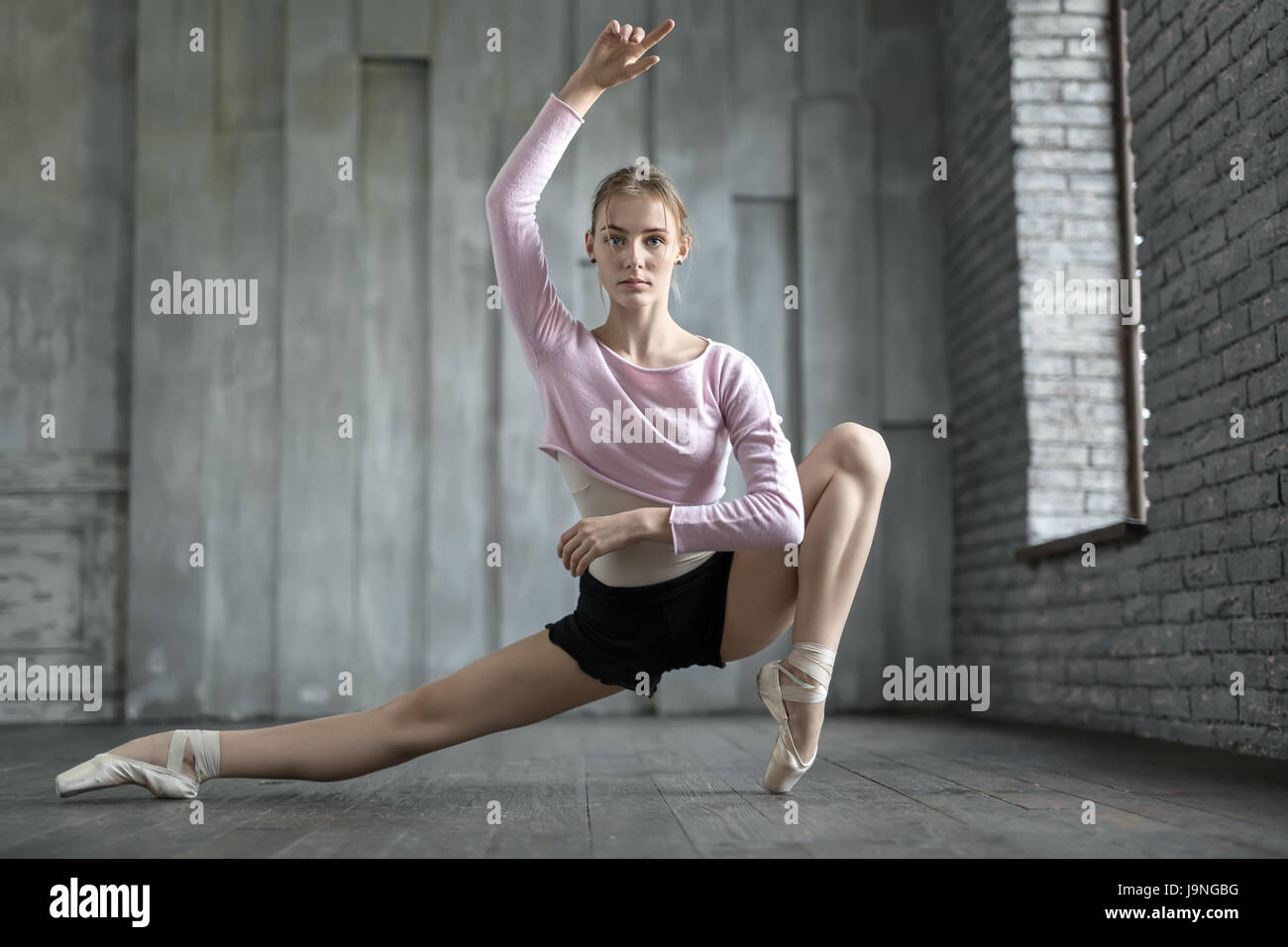 Wunderschöne Ballerina neben dem Fenster auf die graue Wand Hintergrund Hälfte gespalten. Ihr linkes Bein steht auf dem Fuß. Mädchen trägt schwarze Shorts, leichte Trikot Stockfoto