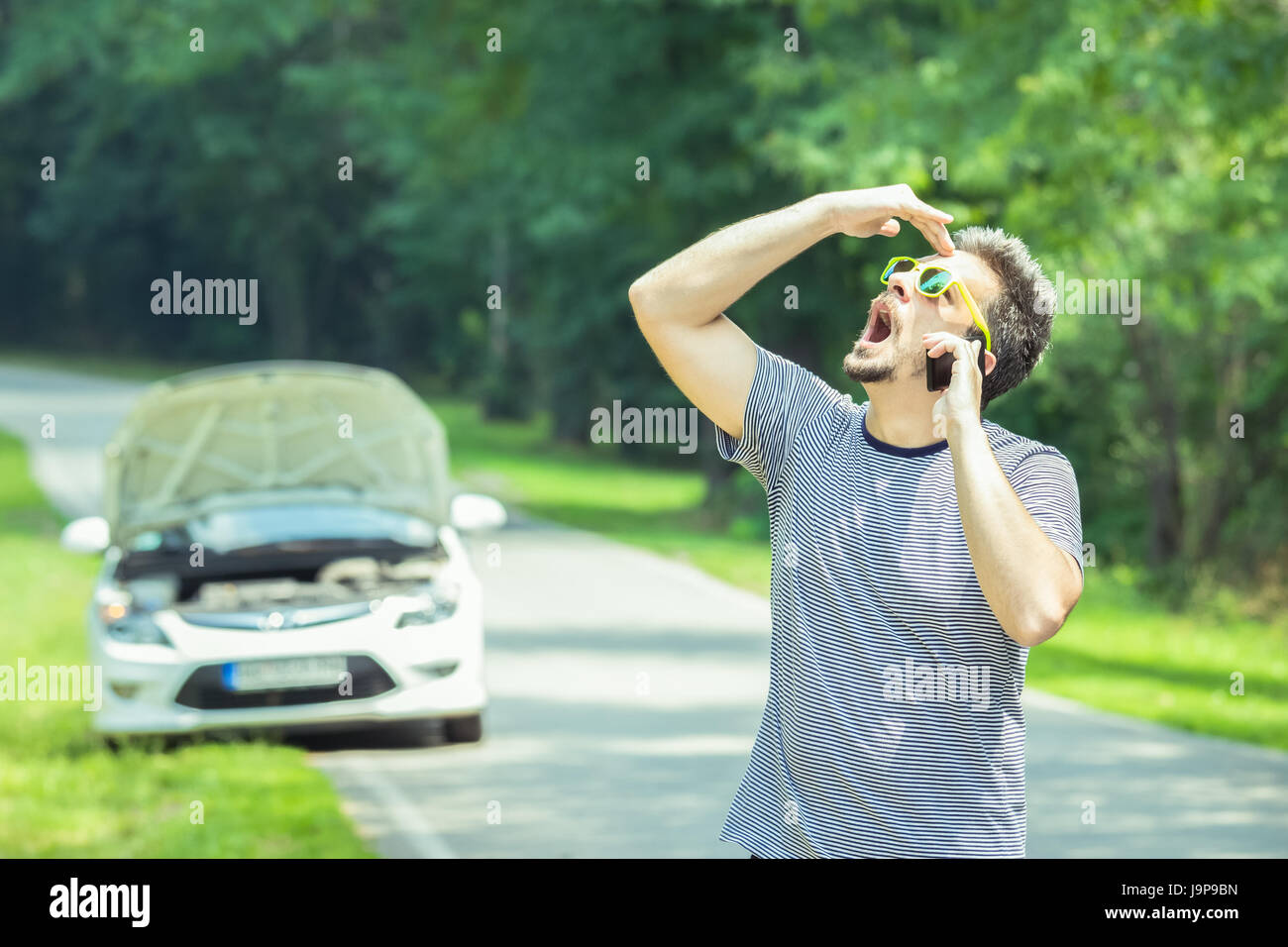 Junge moderne gestresster Mensch machen Anruf mit Hilfe Pannenhilfe mit Smartphone. Auto mit angehobenen Haube am Straßenrand in den Hintergrund. Stockfoto