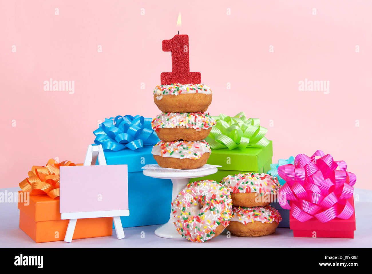 Cake-Donuts mit weißer Glasur und bunten Streuseln gestapelt auf und um ein kleines Podest umgeben von Geburtstagsgeschenken. Nummer 1 Kerze brennen Stockfoto