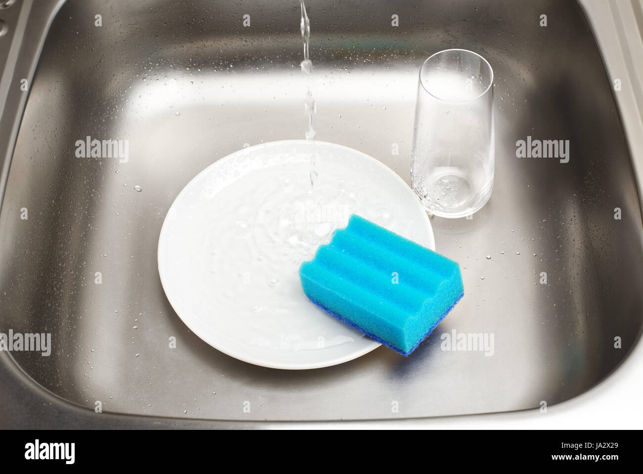 Nahaufnahme der Küchenspüle mit sauberen weißen Teller, blauen Reinigungsschwamm und Glas unter fließendem Leitungswasser trinken Stockfoto