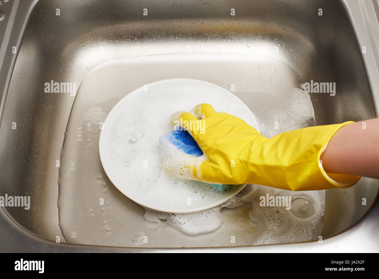 Nahaufnahme von weiblicher Hand in gelben Gummischutz Handschuh waschen weißen Teller mit blauen Reinigungsschwamm gegen Küchenspüle Stockfoto