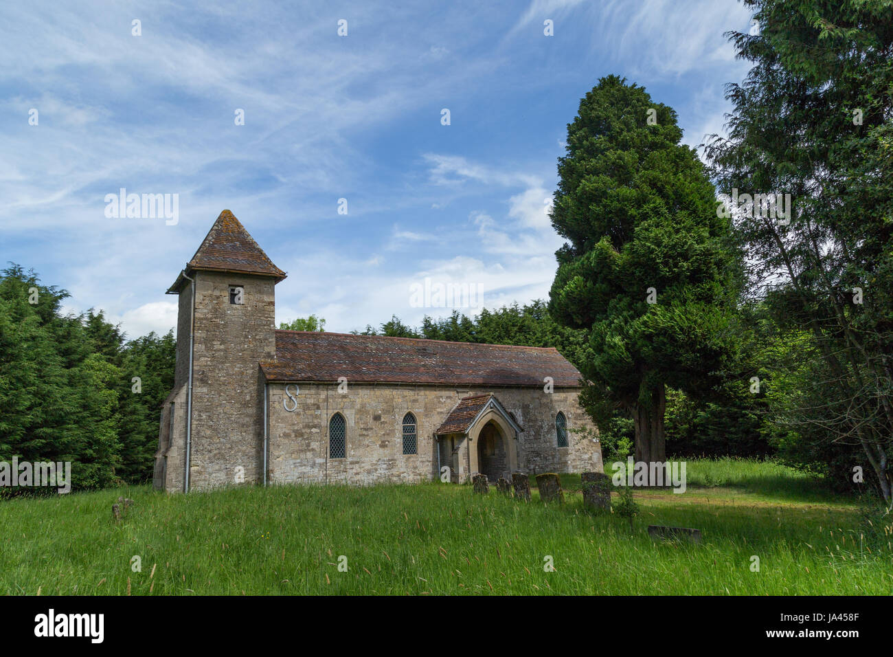 Pfarrkirche Heilige Dreifaltigkeit in Godington in Oxfordshire. Die Kirche ist umgeben von Bäumen auf einer grünen Weide mit blauem Himmel und Wolkenfetzen. Stockfoto