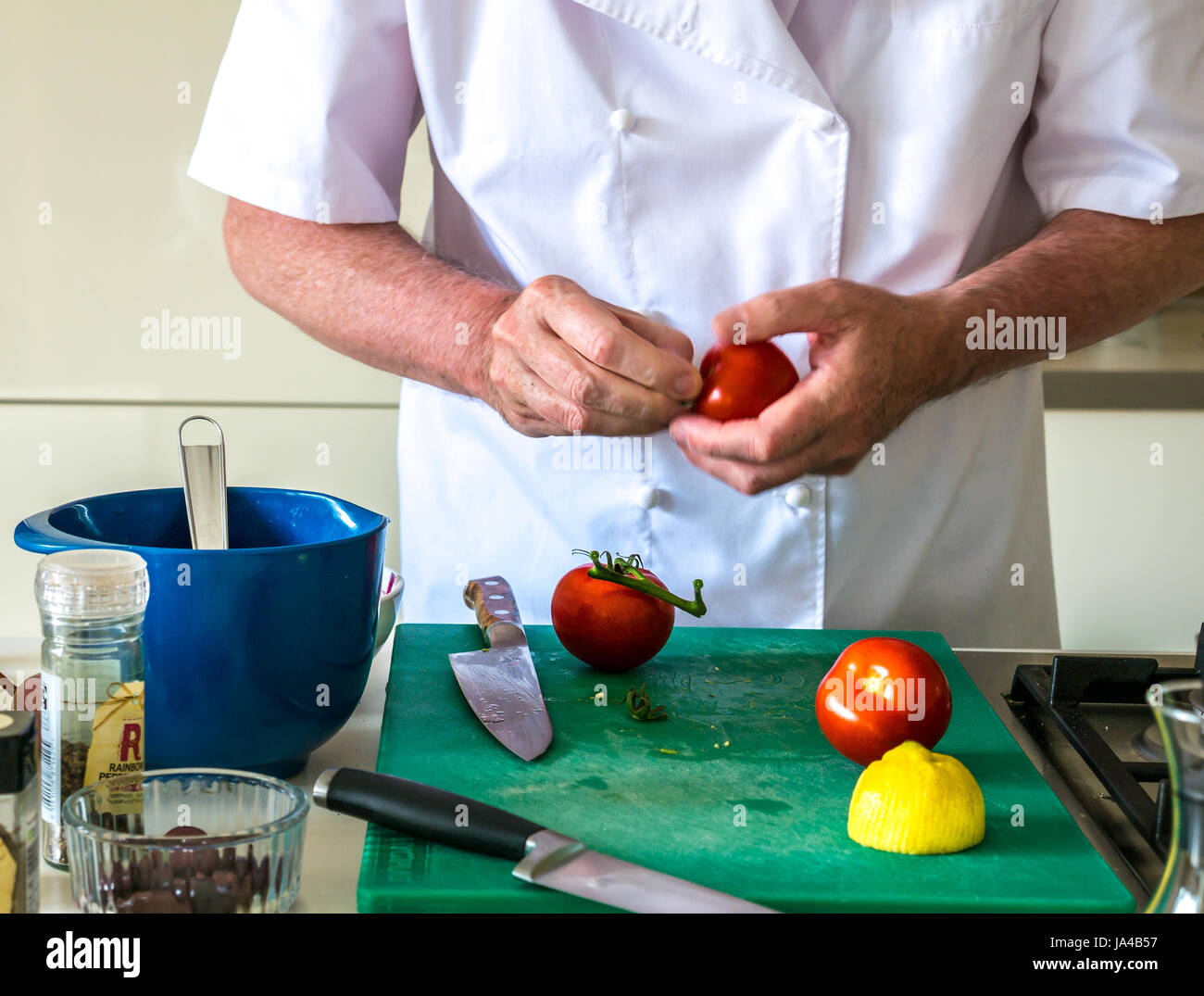 Ein Mann in Chef weiß Zubereitung Tomaten auf einem grünen Schneidebrett auf einem Küchentisch mit anderen Küchengeräten und weißen Schränke im Hintergrund Stockfoto