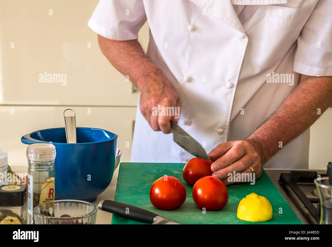 Ältere Mann in Chef weiß Zubereitung Tomaten auf Schneidebrett auf der Arbeitsplatte in der Küche mit Küchengeräten und weißen Schränke im Hintergrund Stockfoto