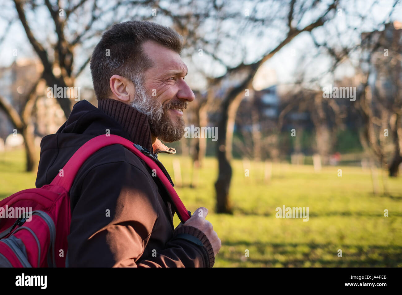 Reifer Mann ist Fuß in einem Park mit einem Rucksack an einem sonnigen Tag und lächelnd. Stockfoto
