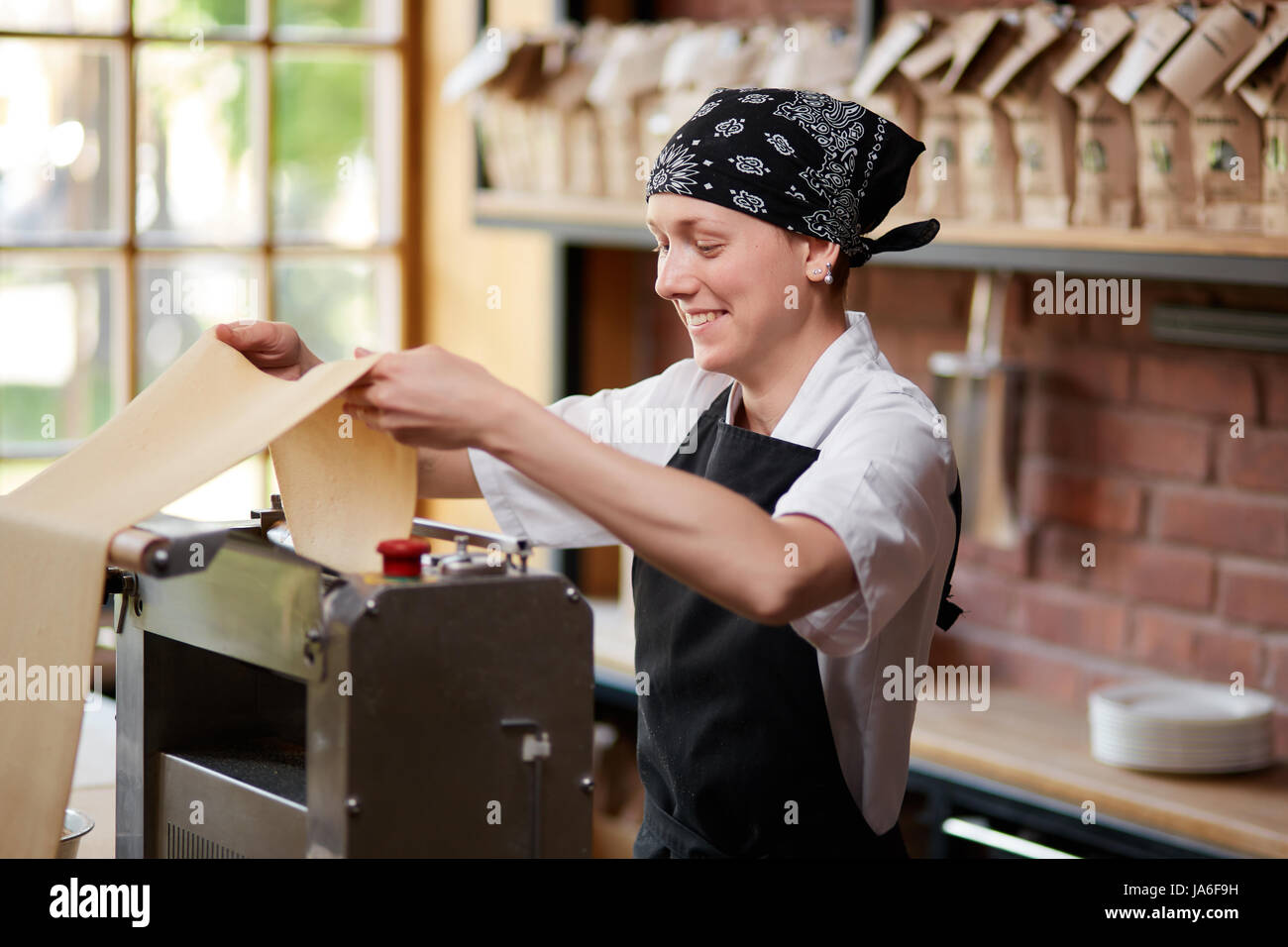 Fröhliche Köchin rollenden Pasta kneten durch Maschine in Cafekithcen. Pastaherstellung. Pastaherstellung im Restaurant. Lächelnde Mitarbeiter Kochen von Teigwaren in r Stockfoto