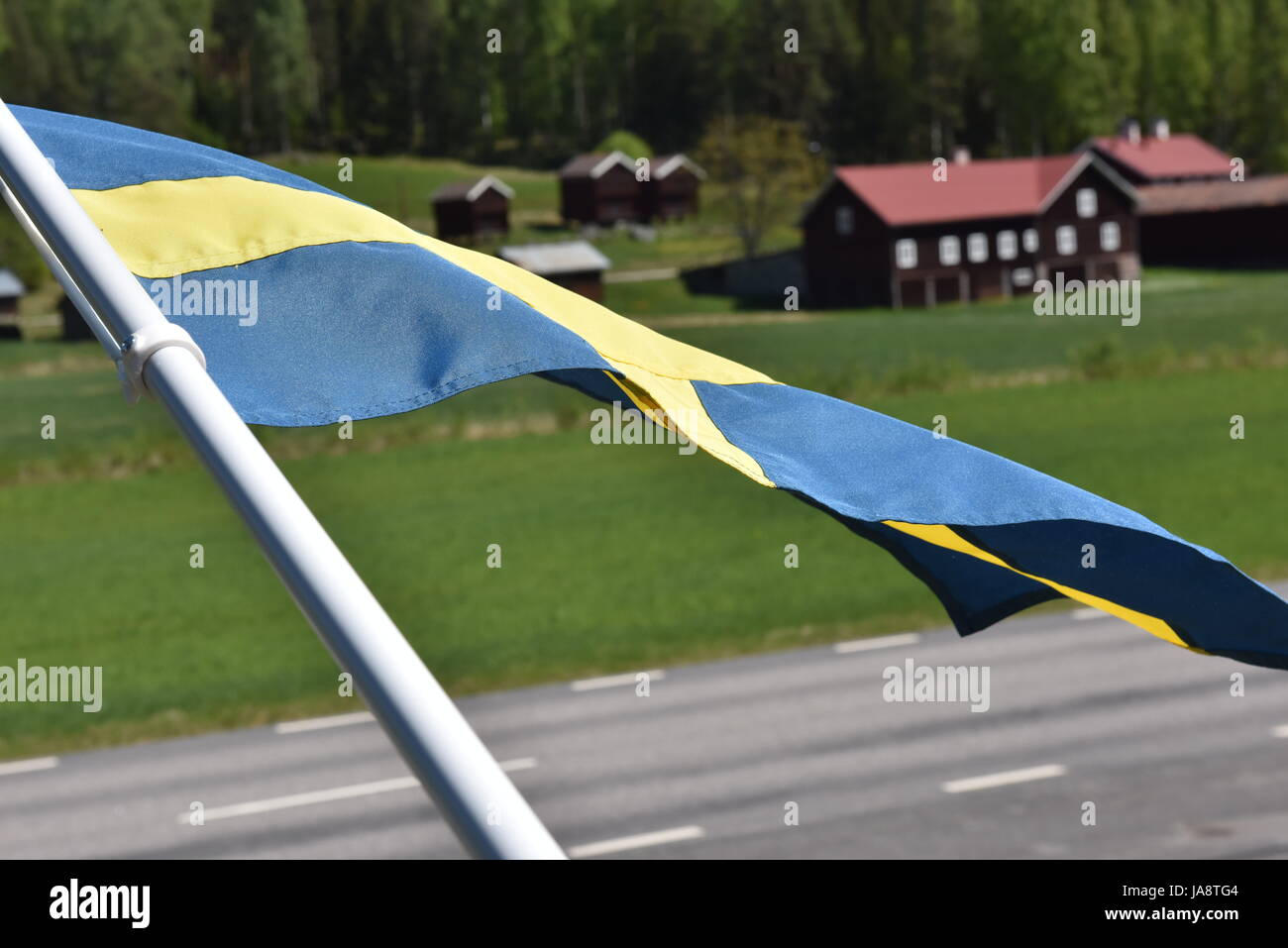 Schwedische Landschaft mit roten Holzhäusern und der schwedischen Flagge, die an einem hellen, sonnigen Sommertag im Vordergrund winkt Stockfoto