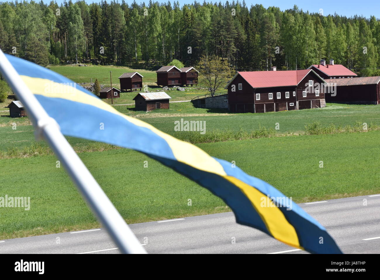 Schwedische Landschaft mit roten Holzhäusern und der schwedischen Flagge, die an einem hellen, sonnigen Sommertag im Vordergrund winkt Stockfoto