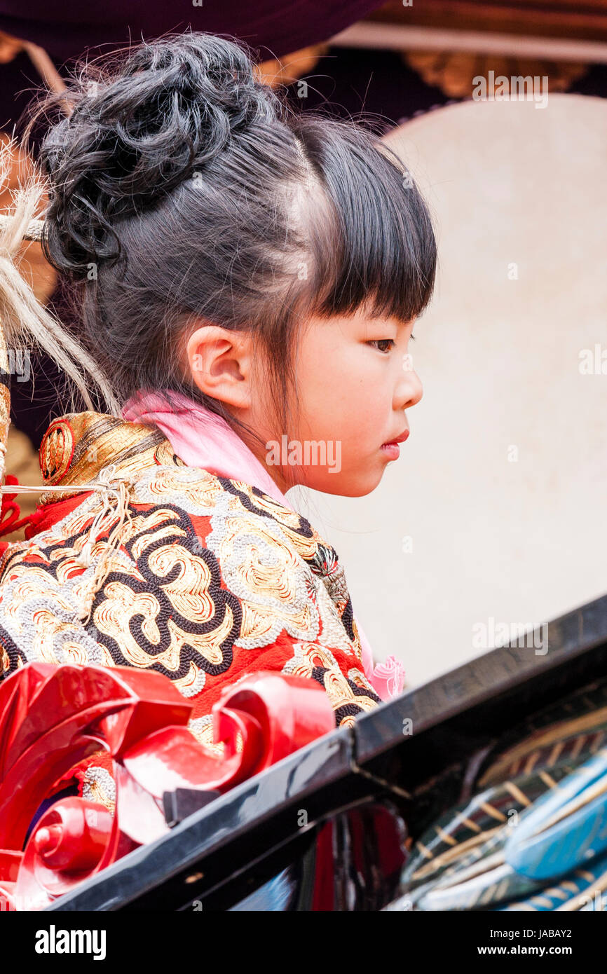 Japanisches Kind, Mädchen, 8-9 Jahre alt, auf der Suche geradeaus sitzend auf Dashi schweben. Von der Seite. Sie trägt einen roten und goldenen Kimono. Stockfoto