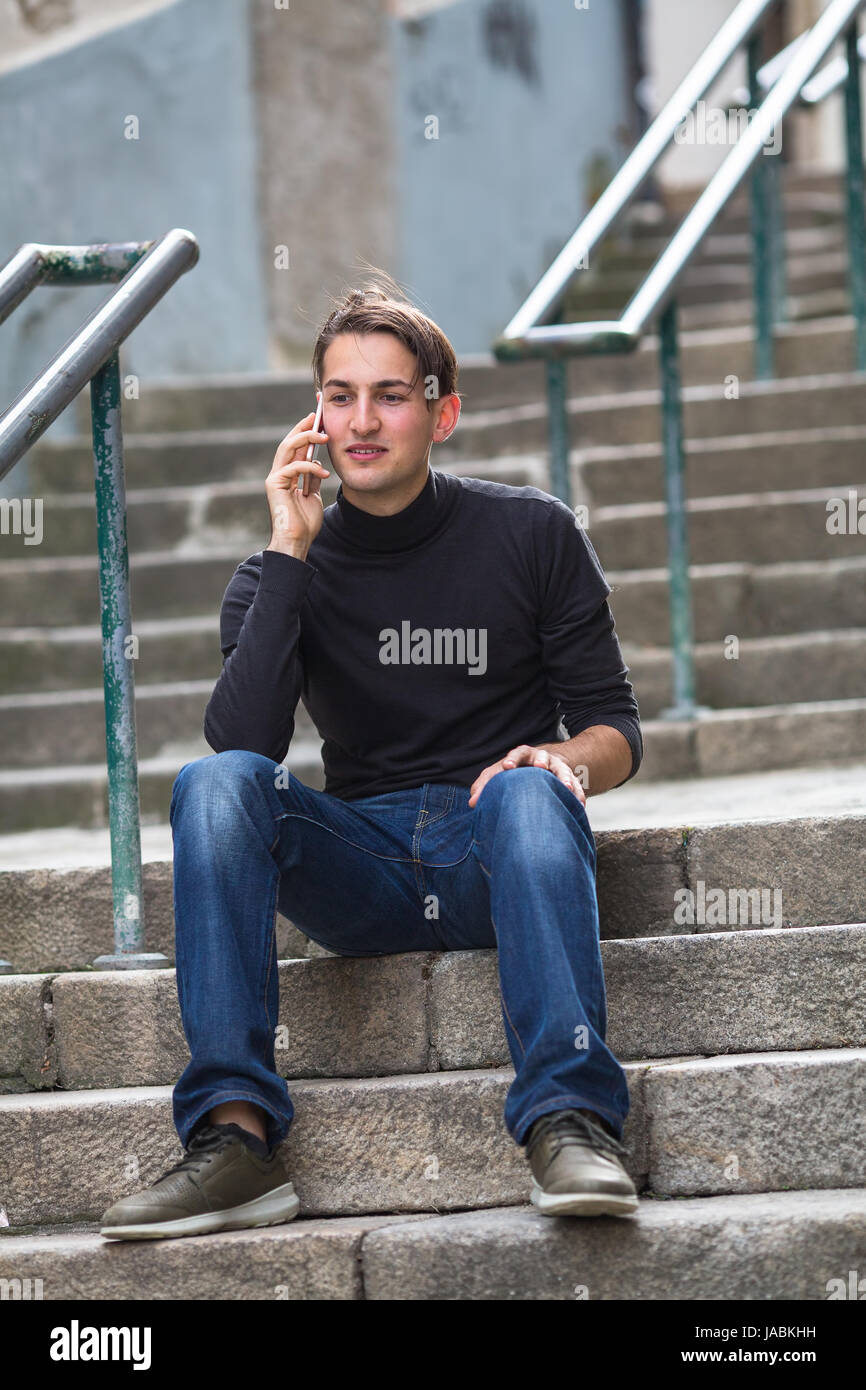 Junger Mann am Telefon sitzt auf der Außentreppe. Stockfoto