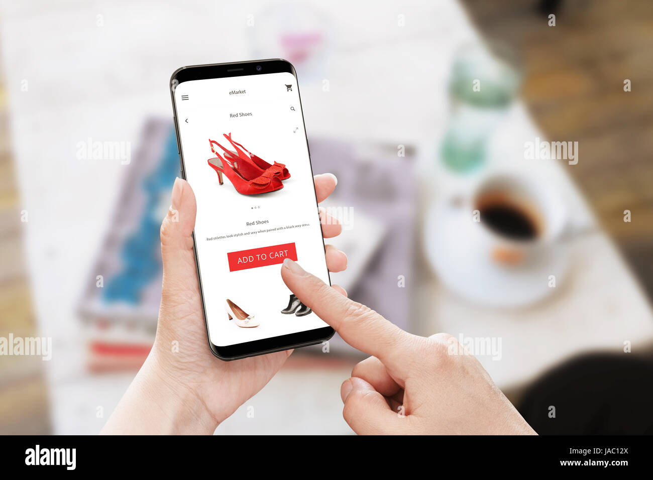 Add to cart rote Schuhe im Online-Shop. Modernes Smartphone mit abgerundeten Ecken in der Hand der Frau. Tisch mit Kaffee und Zeitschriften im Hintergrund. Stockfoto