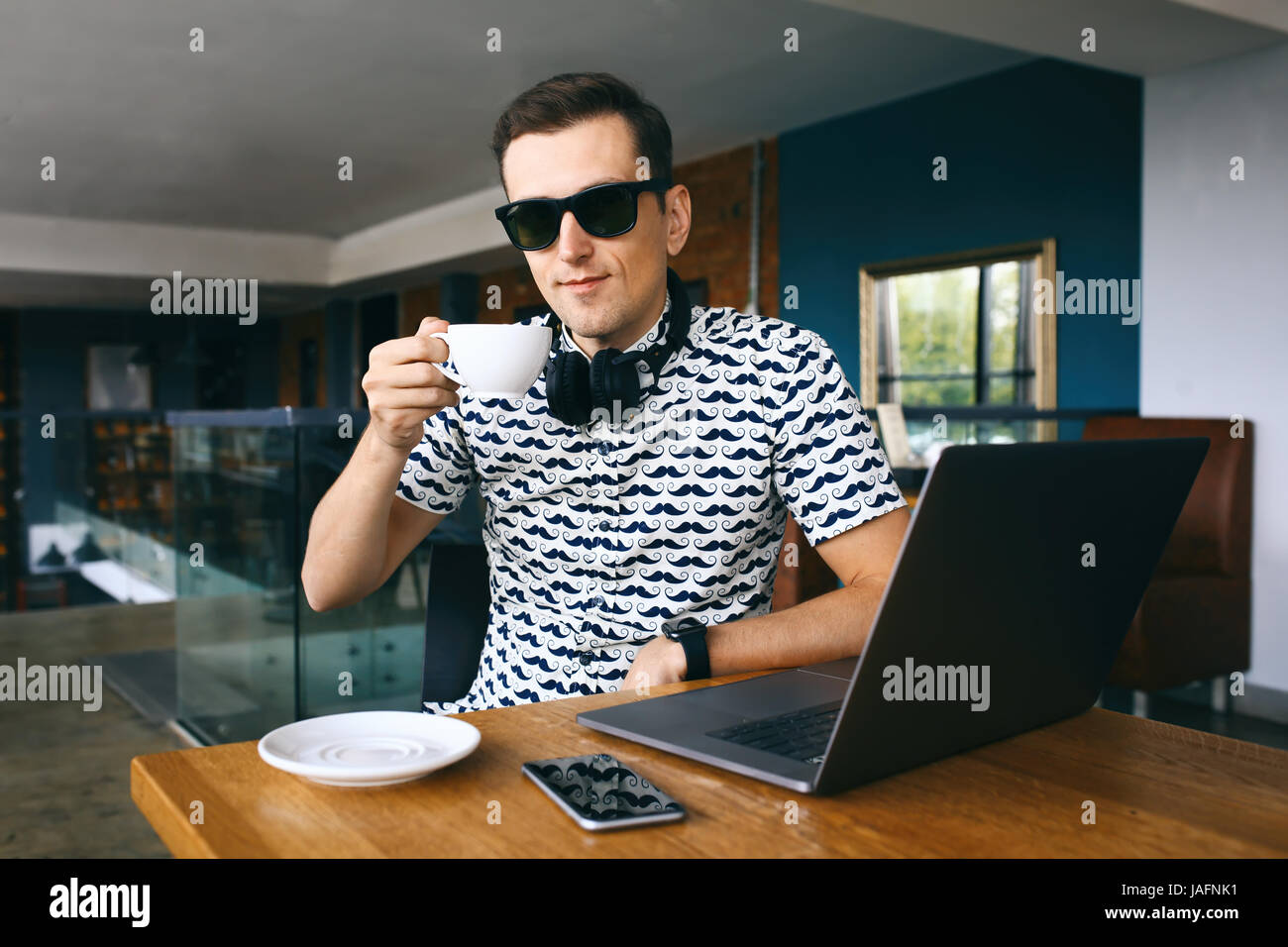 Junge hübsche Hipster Mann sitzt im Café Tasse Kaffee halten. Laptop und Handy auf Holztisch. Stockfoto