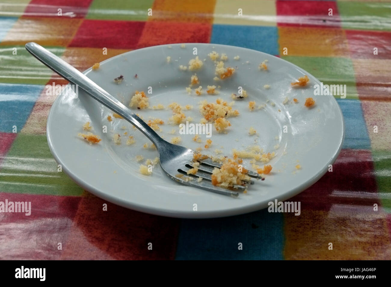 Eine leere kleine Teller mit einer Gabel und die Krümel aus eine Torte, die gegessen hat, sitzt auf einer glänzenden Tischdecke mit Muli-farbige Quadrate. Stockfoto