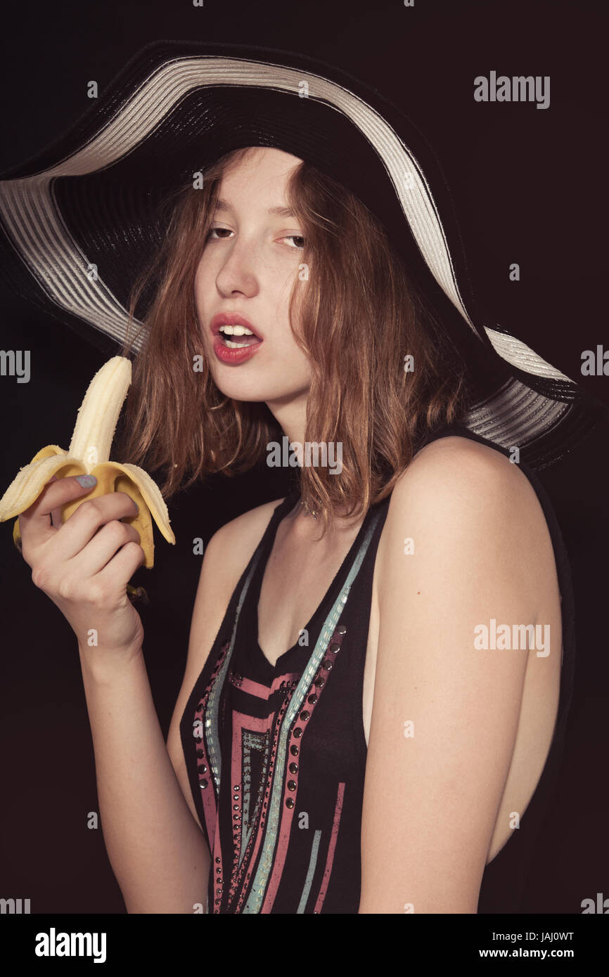 Gekleidet in Sommerhut Mädchen isst eine Banane. Studio Foto auf schwarzem Hintergrund Stockfoto