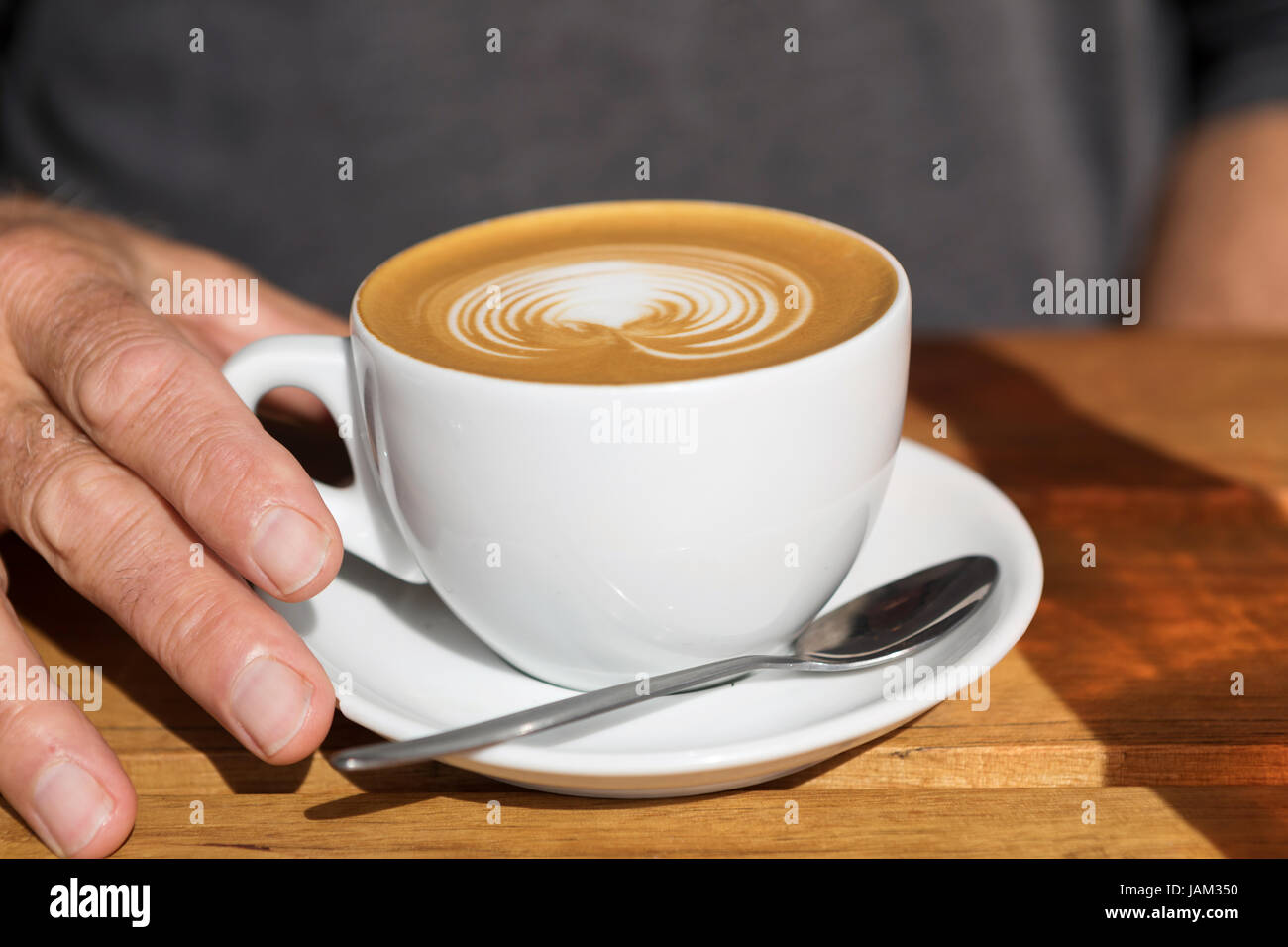 die Hand des Mannes auf dem Griff einer Tasse Latte mit einem Design und einem Löffel auf den Teller Stockfoto