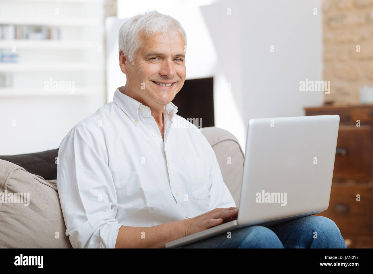 Lächelnd reifen Mann sitzt auf einem Sofa Eingabe auf einem Laptop in einem Wohnzimmer Stockfoto