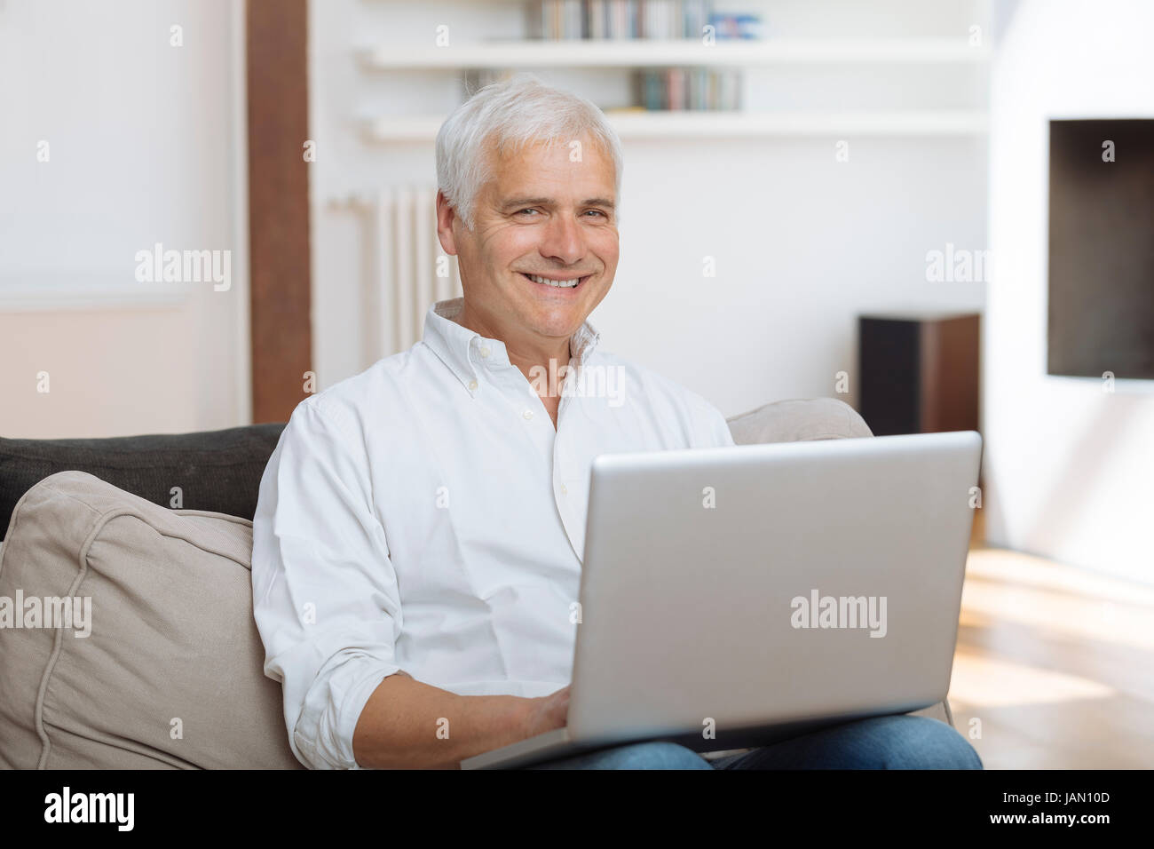 Lächelnd reifen Mann sitzt auf einem Sofa Eingabe auf einem Laptop in einem Wohnzimmer Stockfoto
