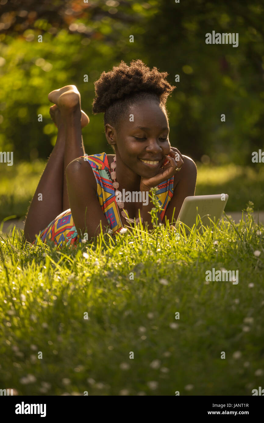 eine, junge Erwachsene, schwarze afroamerikanische glücklich lächelnd genießen Frau 20-29 Jahren, Verlegung auf Rasen lesen Tablet, Natur Park Natur, sonnige d liegend Stockfoto
