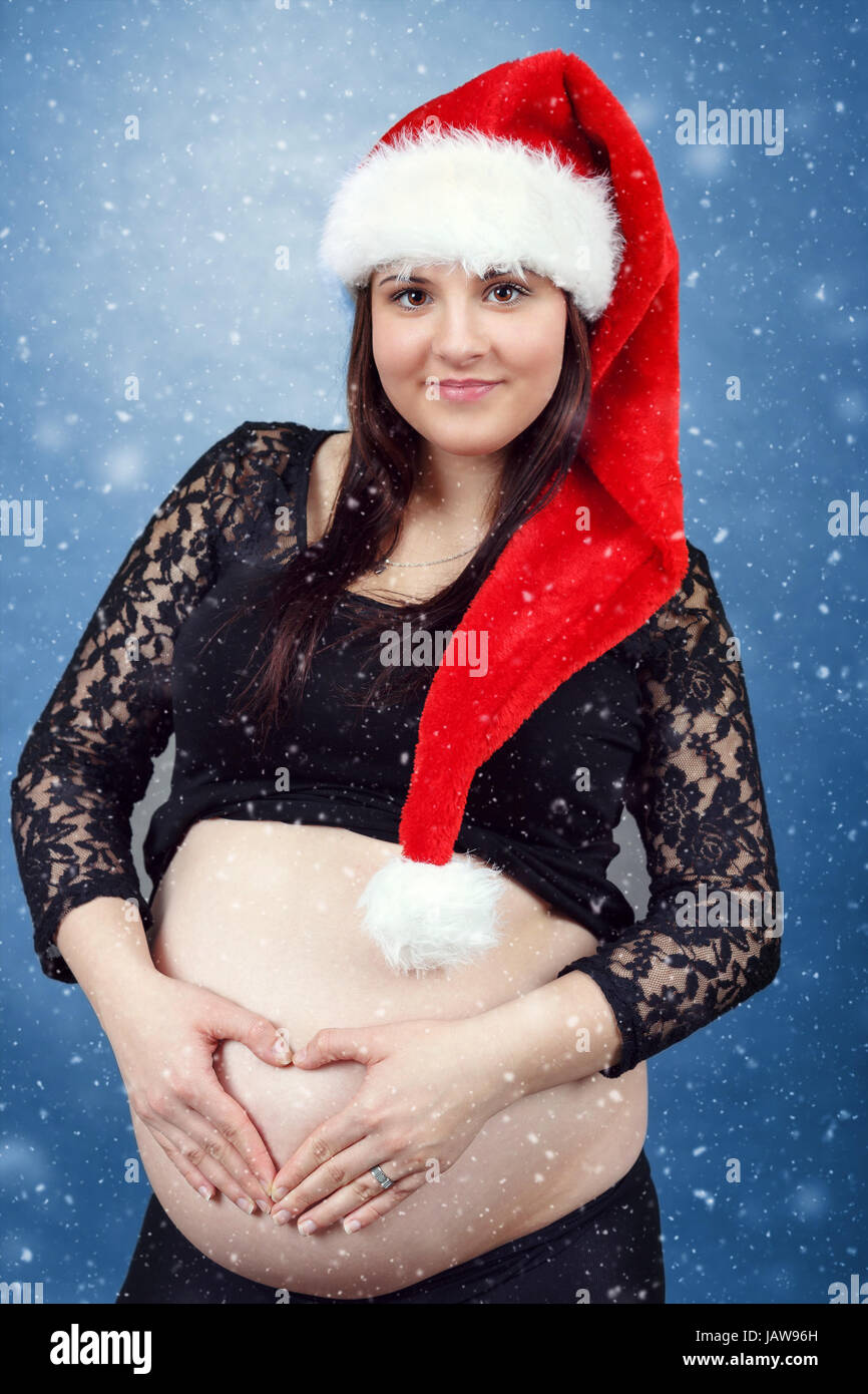 schönen lächelnden jungen Santa schwangere zärtlich hält ihren Bauch, Hand in Herzform und mit Schnee im Hintergrund Stockfoto