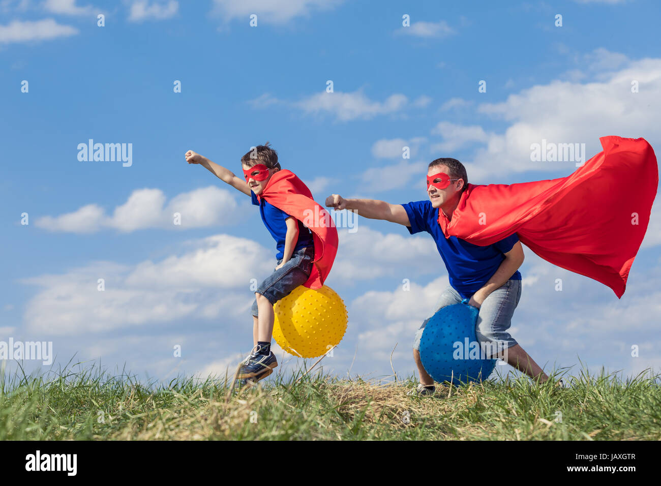 Vater und Sohn spielen Superhelden in der Tageszeit. Leute, die Spaß im Freien. Sie springen auf aufblasbaren Bällen auf dem Rasen. Konzept der freundlichen Familie. Stockfoto