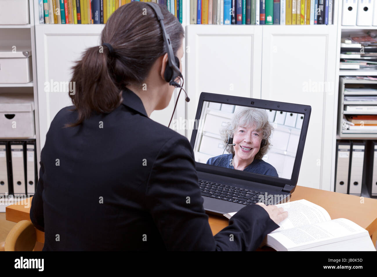 Frau mit Headset vor Ihrem Laptop und ein Buch, einen on-line-Video mit einer älteren weiblichen Kunden oder Auftraggeber, Text Chat Raum Stockfoto