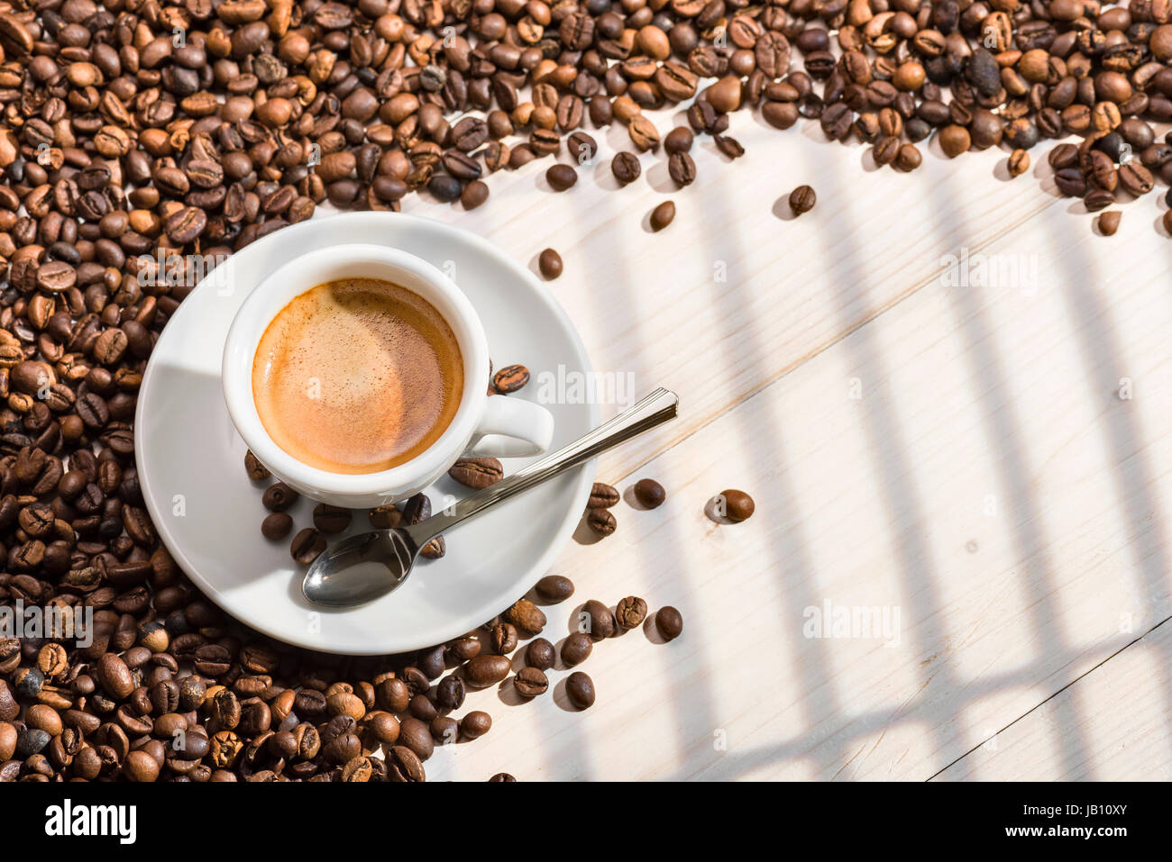 Tasse Kaffee mit Kaffeebohnen auf Holztisch, beleuchtet durch Sonnenlicht, das durch die Fenster. Stockfoto