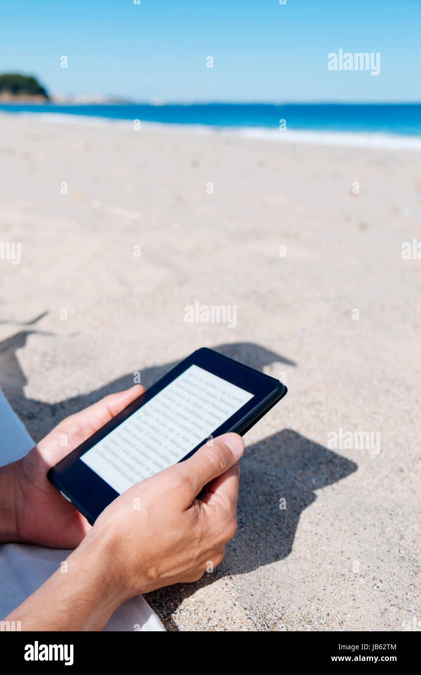 Closeup von einem kaukasischen Jüngling lesen in einem Tablet oder e-Reader auf einem weißen Sandstrand direkt am Meer Stockfoto
