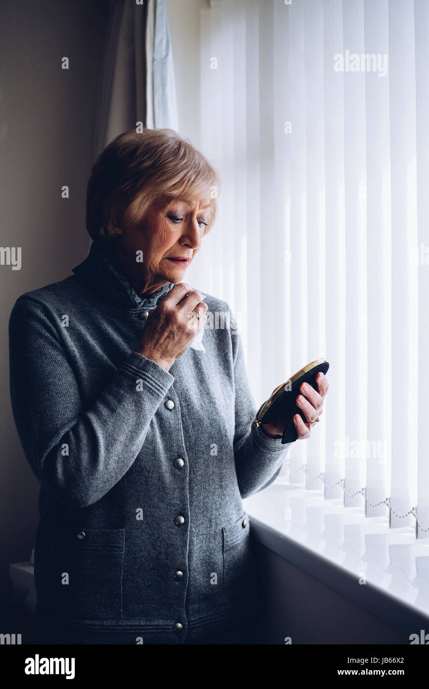 Ältere Frau, die in ihrem Schlafzimmer. In der anderen Hand hält sie ein gerahmtes Foto mit einem Tuch ab. Sie hat einen traurigen Ausdruck auf ihrem Gesicht. Stockfoto