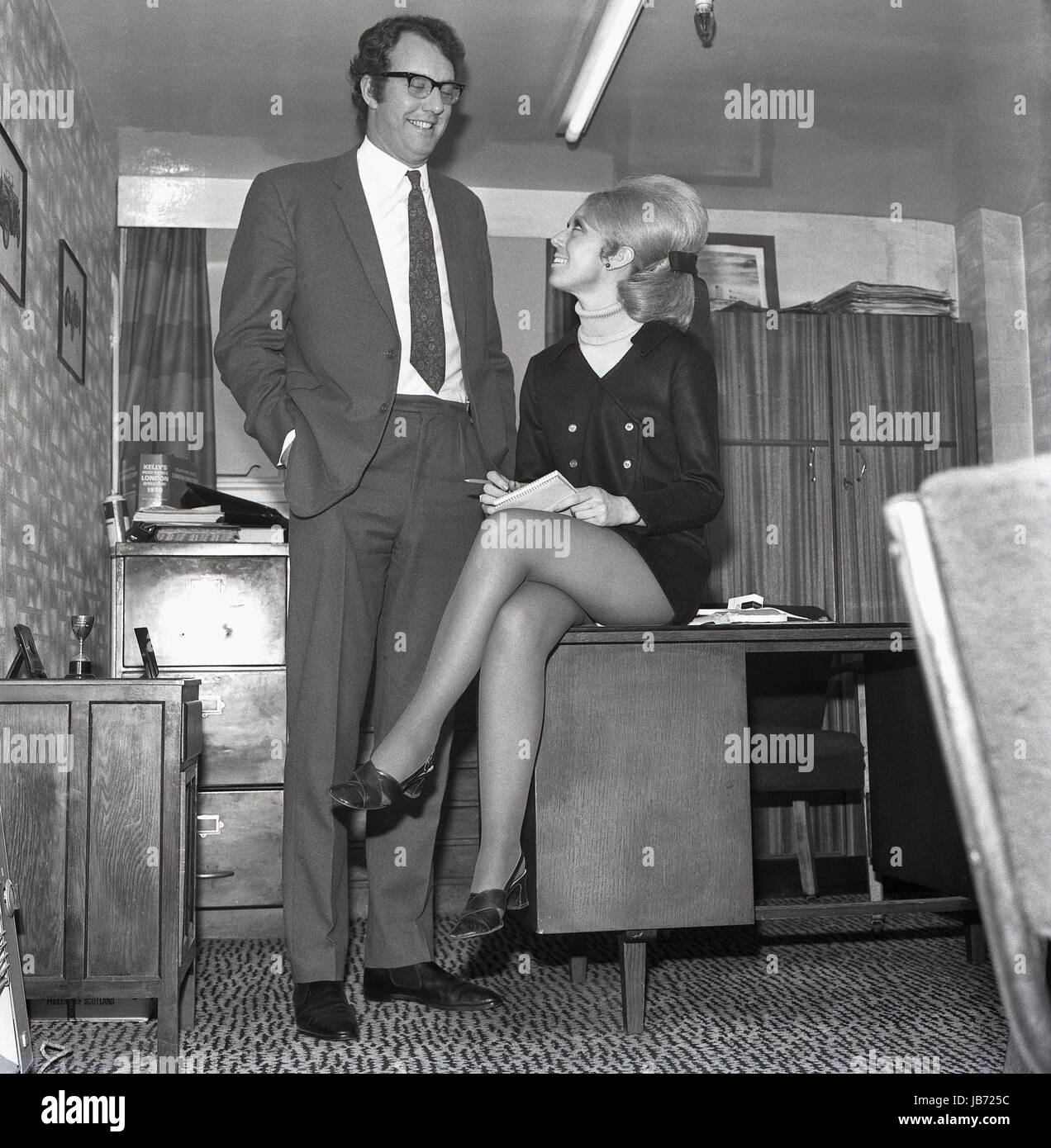 1970er Jahre, historische, trägt einen Minirock Sekretärin sitzt auf dem Rand von einem ihr Chef Schreibtisch als sie Notizen in seinem Büro nimmt. Stockfoto