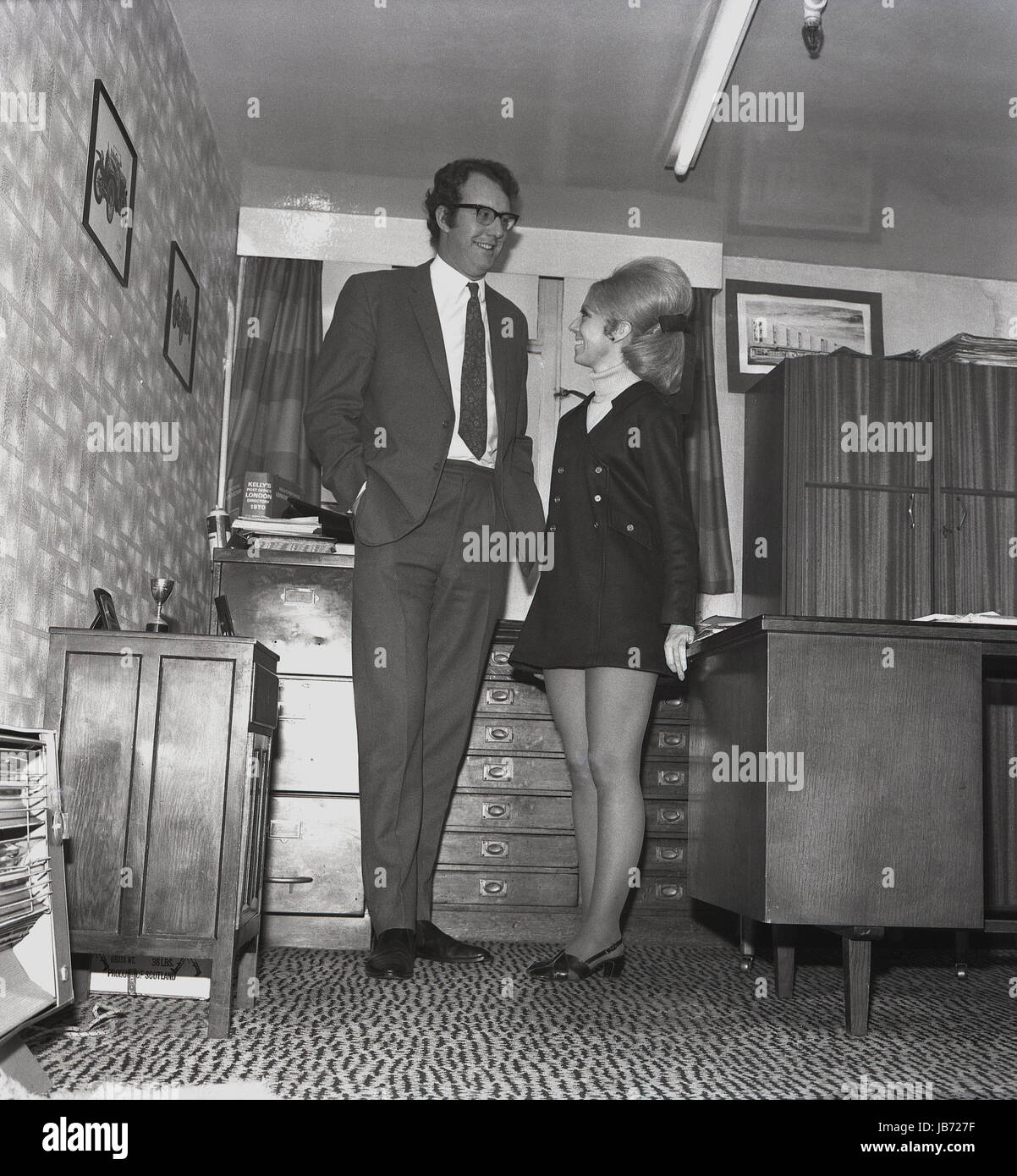 1970er-Jahren, historische, eine Sekretärin trägt einen Mini-Rock und mit einem Buoffant Frisur steht neben ihrem hohen männlichen Chef in seinem Büro. Stockfoto