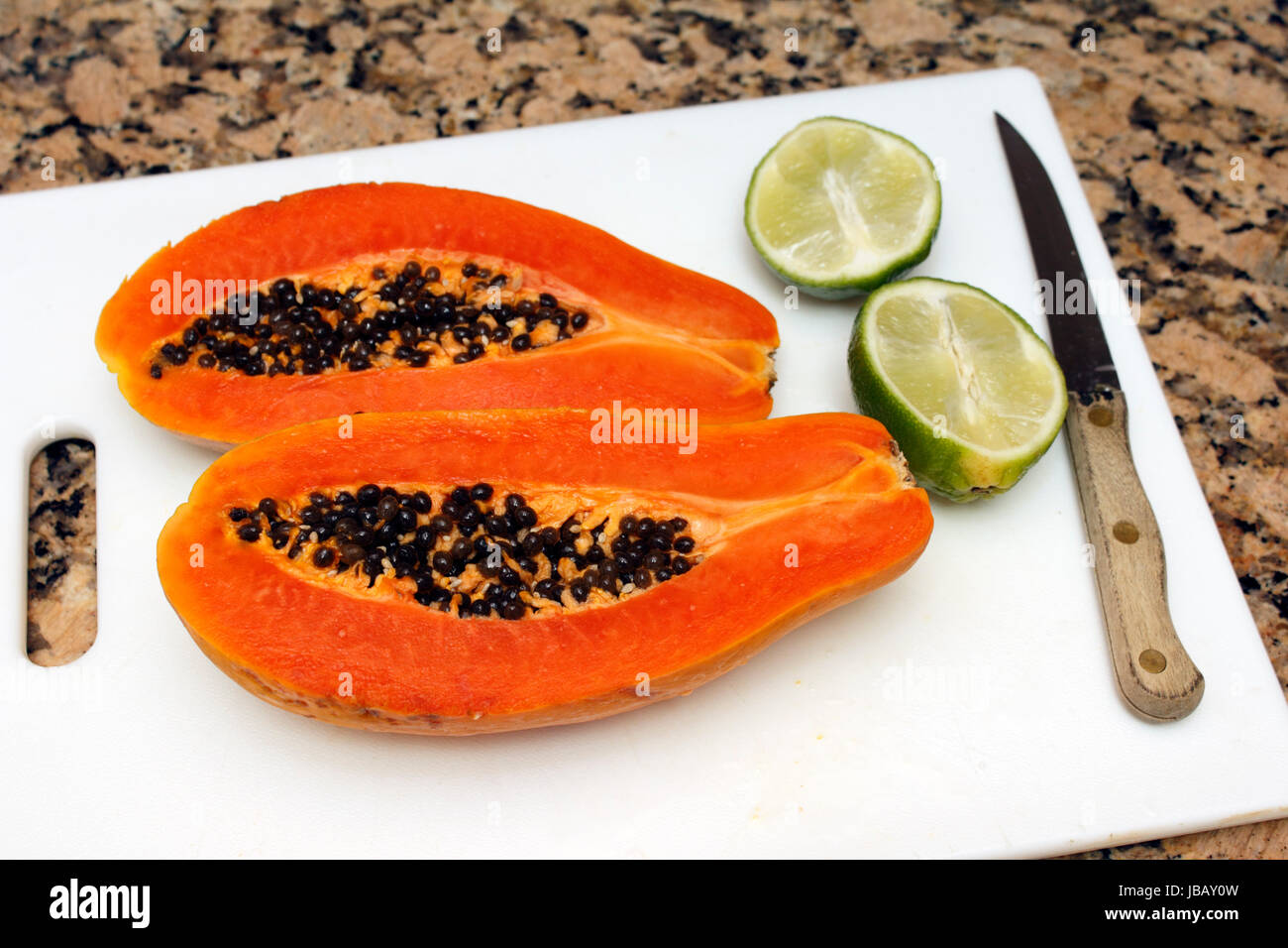 Eine große dunkle orange Papaya Frucht in zwei Hälften geschnitten längs mit schwarzen Samen im Inneren neben zwei Hälften einer Limette auf einer weißen Schneidebrett in der Nähe eines Messers auf einem braunen und schwarzen Granit Küche Zähler nach oben. Stockfoto