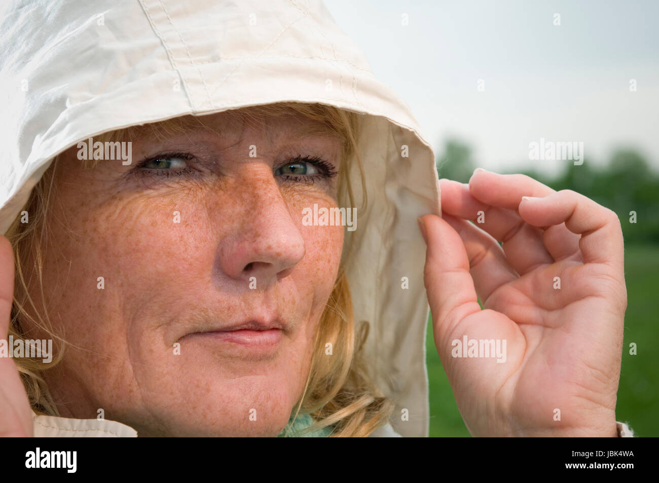 Seitliches Kopf-Porträt Einer blonden Reifen Frau in Heller Jacke Mit Kapuze Auf Einer Grünen Wiese-auflösende in Die Kamera Blickend; Finger der Linken Hand Seitlich eine der Kapuze Stockfoto