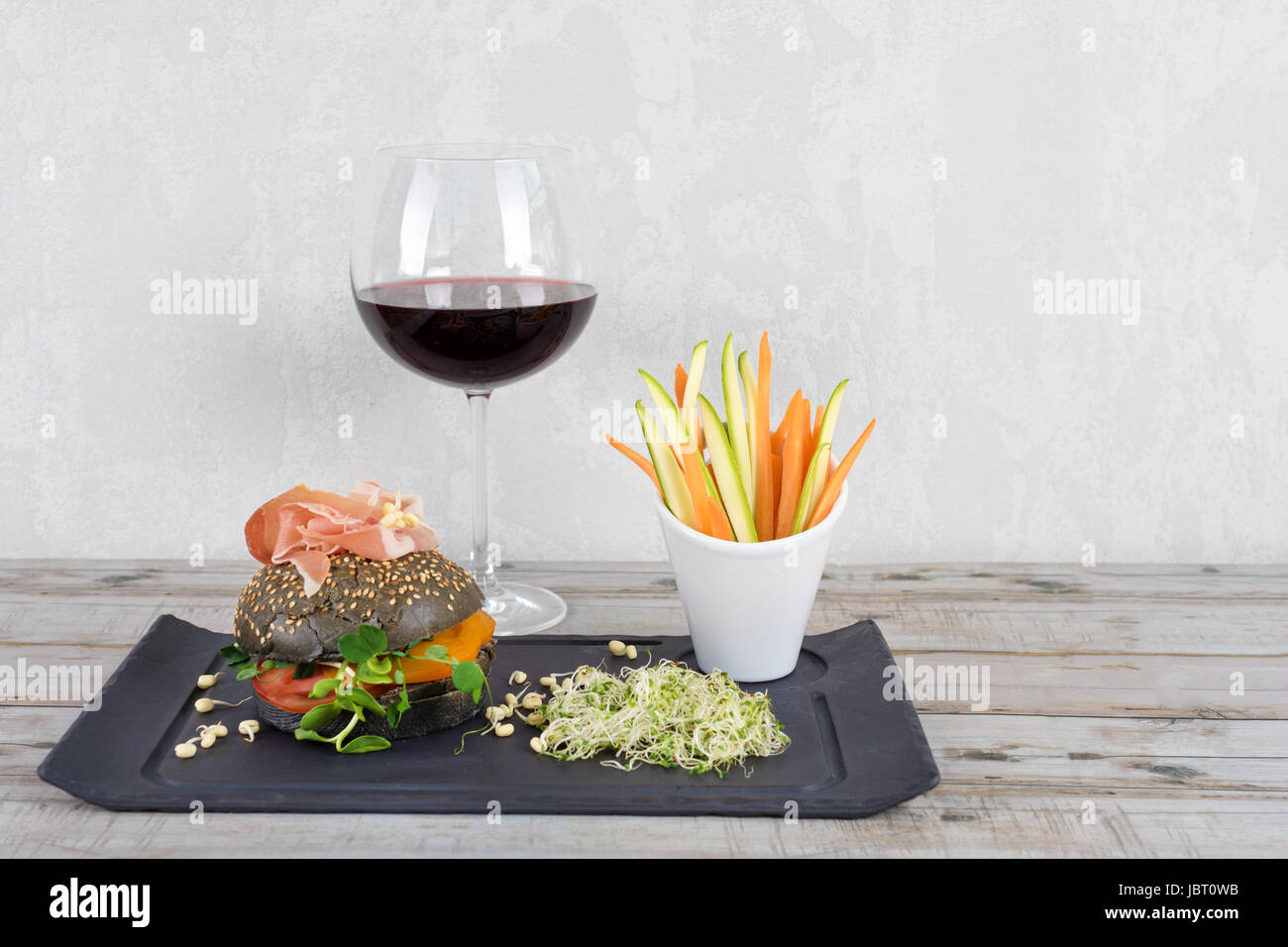 Gesunde Burger mit Hamon, Tomaten, Mikro grünen und schwarzen Vollkorn Brötchen, Gemüsesticks und Rotwein auf Schiefer Tafel über hölzerne Hintergrund. Sauber Essen, Diäten, detox Food-Konzept. Stockfoto