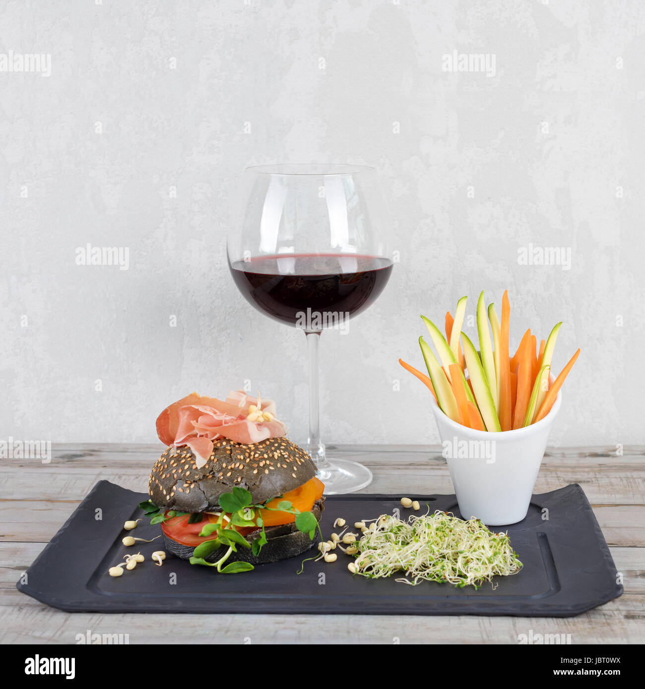 Gesunde Burger mit Hamon, Tomaten, Mikro grünen und schwarzen Vollkorn Brötchen, Gemüsesticks und Rotwein auf Schiefer Tafel über hölzerne Hintergrund. Sauber Essen, Diäten, detox Food-Konzept. Stockfoto