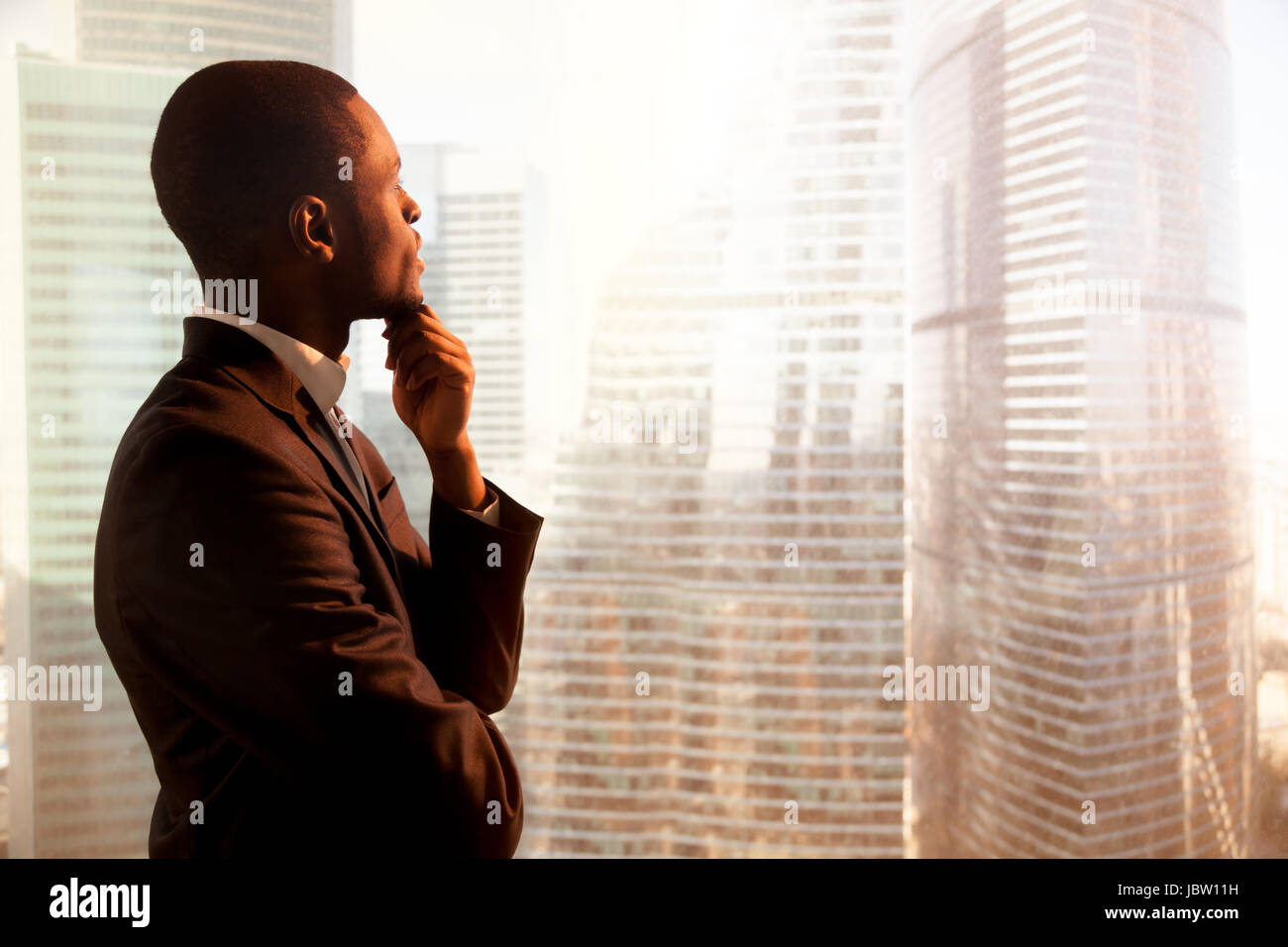 African-American nachdenklich Jungunternehmer Anzug an Hand am Kinn, Blick aus großen Bürofenster auf Sonnenuntergang Stadt Gebäude in du verloren Stockfoto