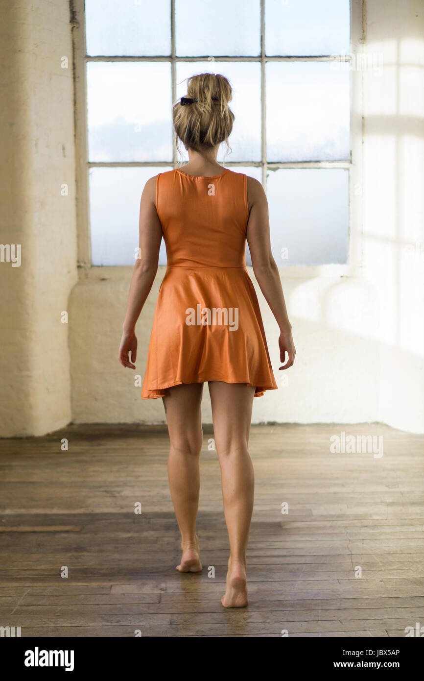 Rückansicht einer jungen blonden kaukasischen Frau trägt ein kurzen orange Sommerkleid zu Fuß in Richtung des Fensters barfuß Stockfoto