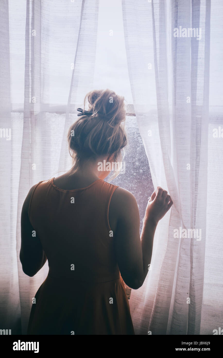 Rückansicht einer blonden kaukasischen Frau steht am Fenster, wegschauen Stockfoto