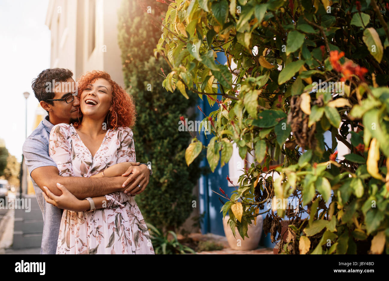 Mann umarmt seine Partnerin von hinten seine Arme um sie wickeln. Lächelnde Frau Hand in Hand eines Mannes romantisch mit ihr Gesicht dicht vor seinem Gesicht. Stockfoto