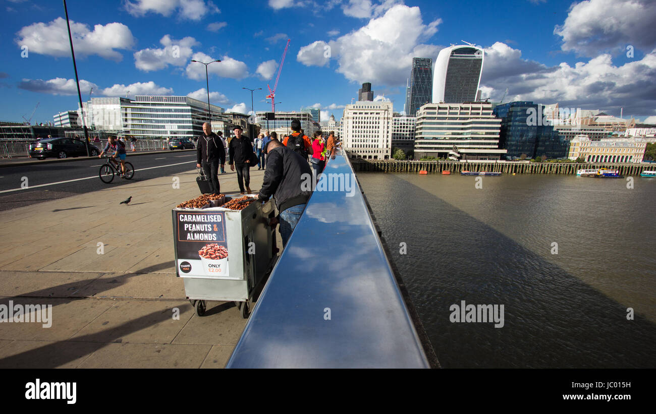 Touristen und einheimische mischen sich auf London Bridge über die Themse in der Sonne unter einem dramatischen Himmel mit jagenden Wolken spiegeln sich in der Schiene Brücke Stockfoto