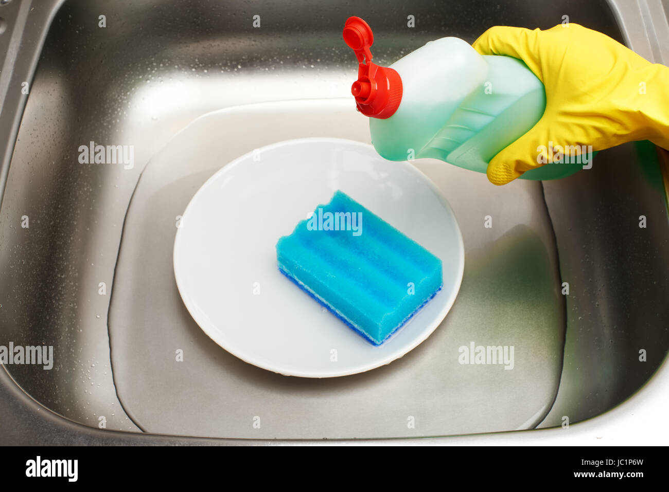 Nahaufnahme der Küchenspüle mit Wasser, saubere weiße Platte, blaue Reinigungs Schwamm und Flasche Spülmittel in weiblicher Hand in gelben Gummischutz Stockfoto