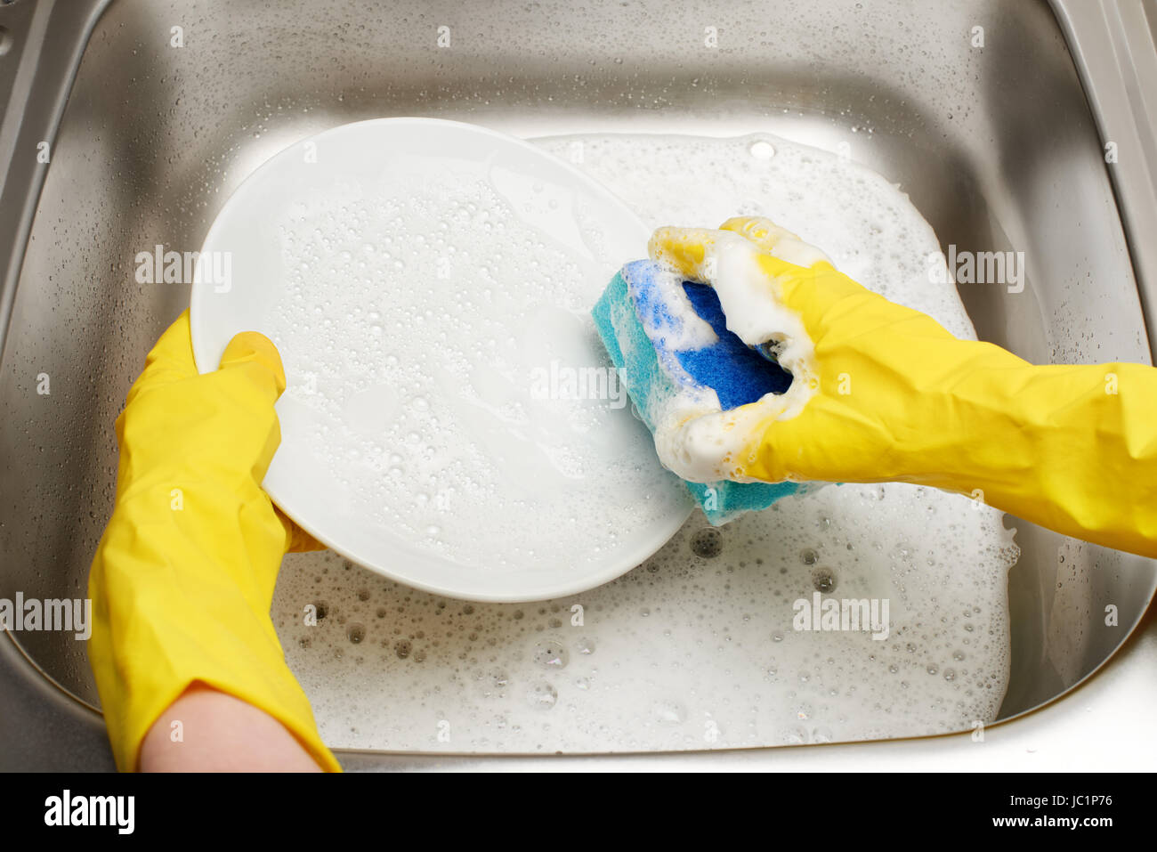 Nahaufnahme von weiblichen Händen Gummischutz Handschuhe waschen weißen Teller mit blauen Reinigung gegen Küchenspüle voller Schaum Schwamm gelb Stockfoto